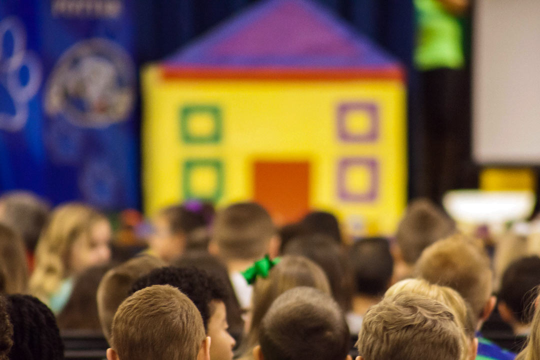 test Rows of elementary school aged children, sitting and facing toward a stage where there is the front of a large yellow house with a purple roof.